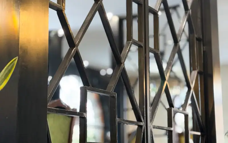 Primer plano de una reja decorativa de una puerta de metal negro con motivos geométricos, incluidos cuadrados y rombos. El fondo está suavemente difuminado con luces y elementos de interior visibles.