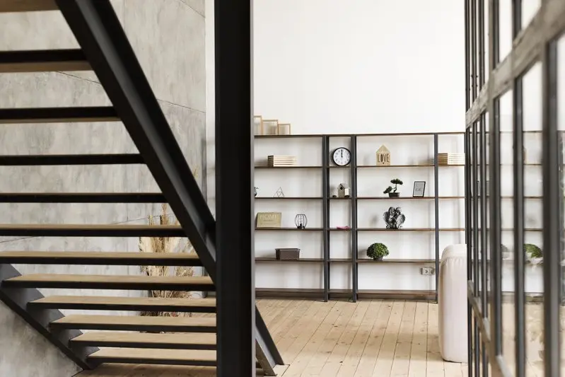 Interior moderno con una escalera de madera abierta, suelo de madera clara y una estantería minimalista contra una pared blanca, donde se exponen libros, plantas, un reloj y artículos de decoración. Los grandes ventanales dejan entrar la luz natural.