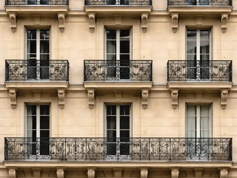 Fachada simétrica de un edificio de piedra beige de tres plantas, cada una de ellas con puertas francesas y balcones ornamentados de hierro forjado negro. Las ventanas son altas y rectangulares, con algunas cortinas visibles en el interior.