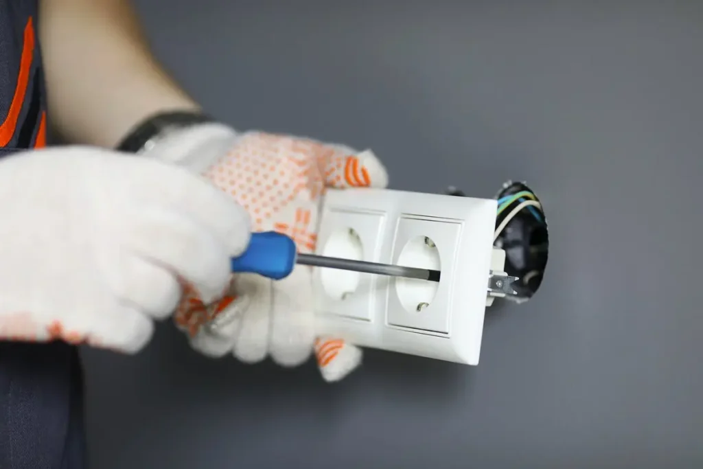 Una persona con guantes de trabajo blancos utiliza un destornillador para instalar o reparar un enchufe doble de pared, con los cables expuestos visibles desde la pared.