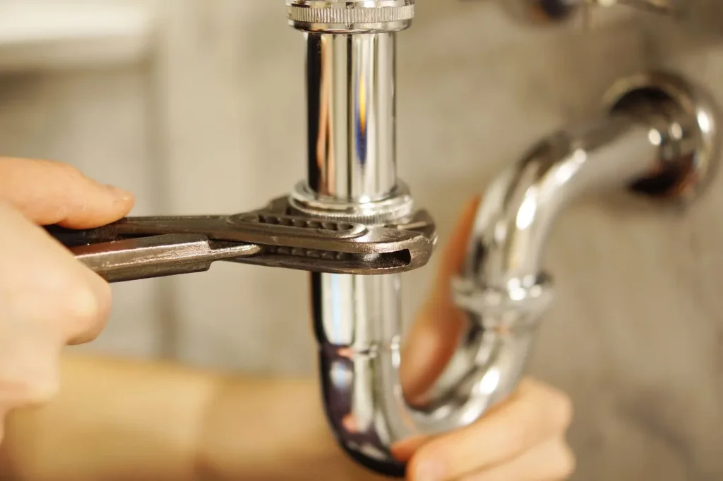 Un fontanero profesional utiliza una llave inglesa para apretar o aflojar un tubo de metal brillante situado bajo un fregadero, lo que indica que se están realizando trabajos de reparación o mantenimiento de fontanería.