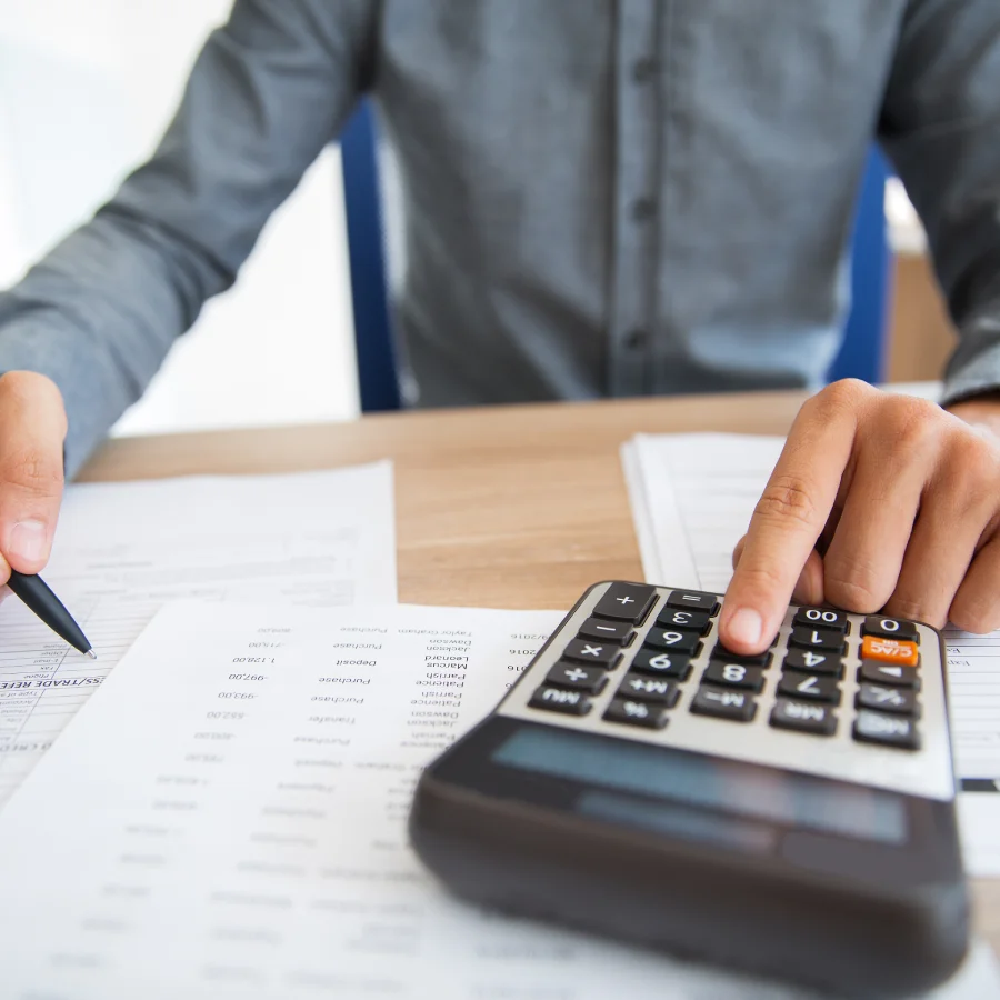 Una persona con camisa gris utiliza una calculadora y sujeta un bolígrafo mientras trabaja con documentos y hojas de cálculo sobre un escritorio.